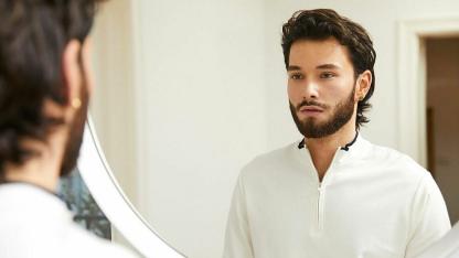 Ein junger Mensch mit Bart schaut in den Spiegel